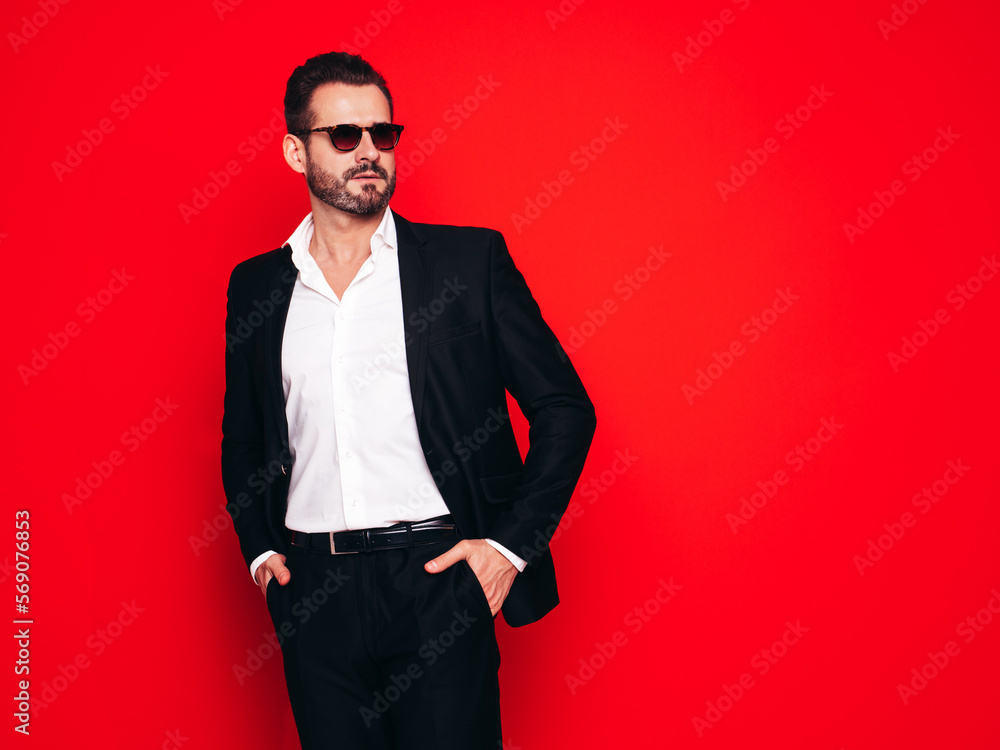 Portrait of handsome confident stylish hipster lambersexual model. Sexy modern man dressed in black elegant suit. Fashion male posing in studio, isolated on red. In sunglasses Portrait of handsome confident stylish hipster lambersexual model. Sexy modern man dressed in black elegant suit. Fashion male posing in studio, isolated on red. In sunglasses