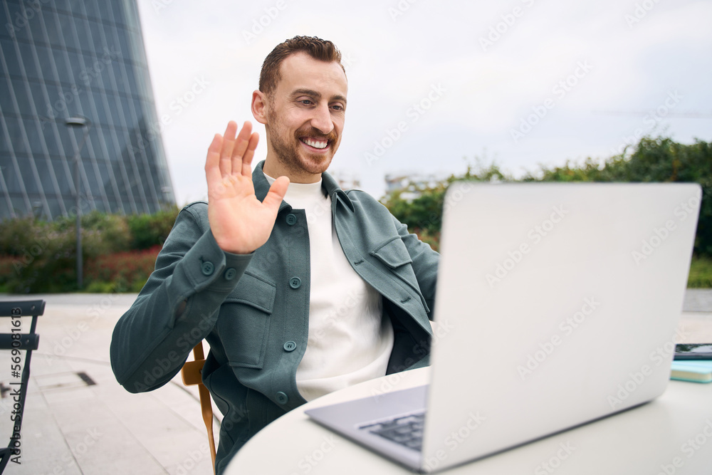 Handsome smiling modern man waving hand to laptop web camera while chatting with family online at street cafe. Video call concept Handsome smiling modern man waving hand to laptop web camera while chatting with family online at street cafe. Video call concept