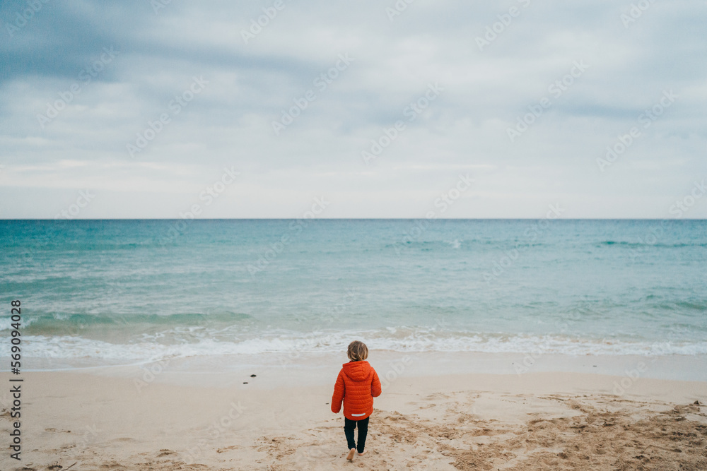 Back view of child wearing jacket walking to sea on cloudy day Back view of child wearing jacket walking to sea on cloudy day