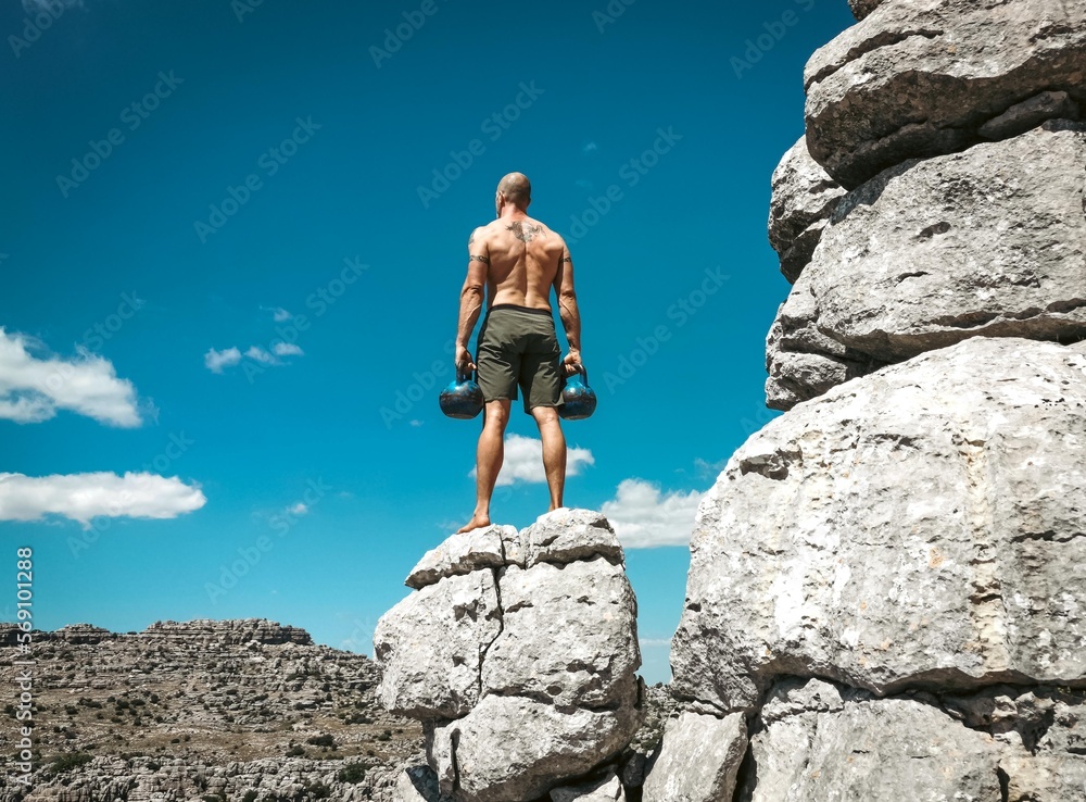 Muscular white male in shorts holding two kettlebells while standing on an ancient rock formation in El Torcal Spain.