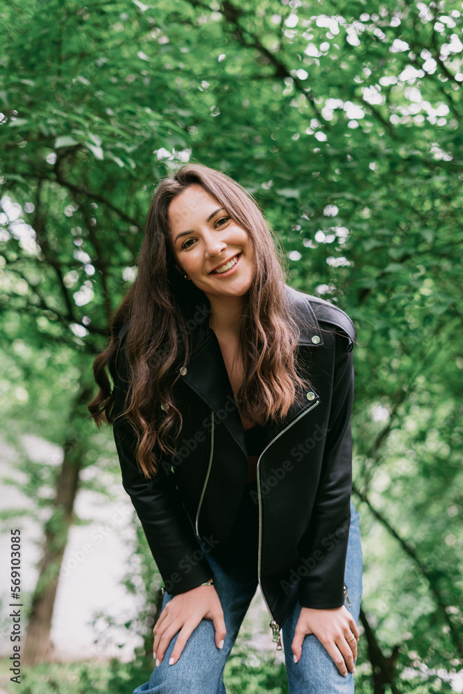 A young woman in a leather jacket poses in the park. Happy smiling model A young woman in a leather jacket poses in the park. Happy smiling model