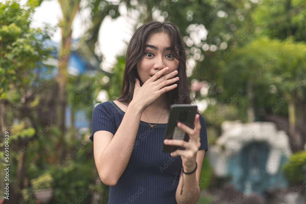 A simple young asian woman shocked, remembering something while holding a phone. Wearing a dark blue blouse. Outdoor garden scene.