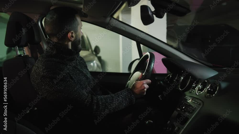 Man in sweater getting in his car and putting on the safety belt. Brunette driver checking rear view mirror and pushing button to start the engine. Bearded guy in auto leaving parking lot