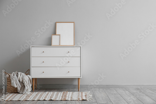 White chest of drawers with blank frames and basket near grey wall © Pixel-Shot