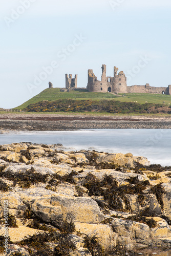 Long exposure photography of the shore and rocky beach between the villages of Craster and Embleton on the Northumberland coast, UK, against background of Dunstanburgh Castle ruins