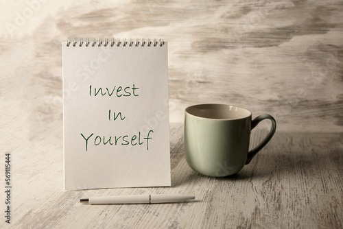 The inscription invest in yourself on a notebook and a cup on the table.