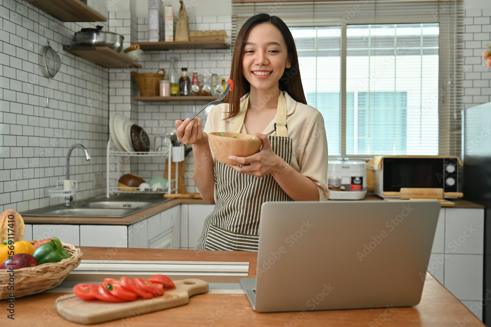 Attractive woman holding salad bowl and making video call on laptop in kitchen at home Attractive woman holding salad bowl and making video call on laptop in kitchen at home
