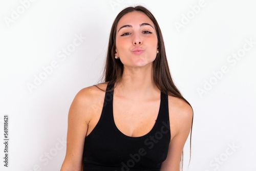 young woman wearing sportswear over white studio background, keeps lips as going to kiss someone, has glad expression, grimace face. Standing indoors. Beauty concept. © Jihan