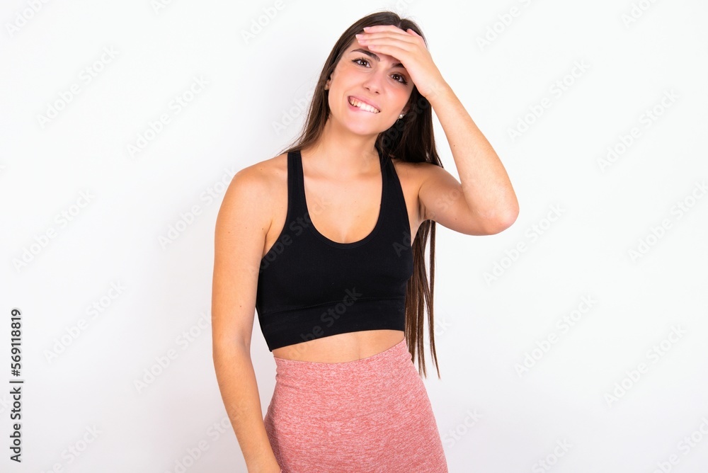 Oops, what did I do? young woman wearing sportswear over white studio background holding hand on forehead with frightened and regret expression. Oops, what did I do? young woman wearing sportswear over white studio background holding hand on forehead with frightened and regret expression.