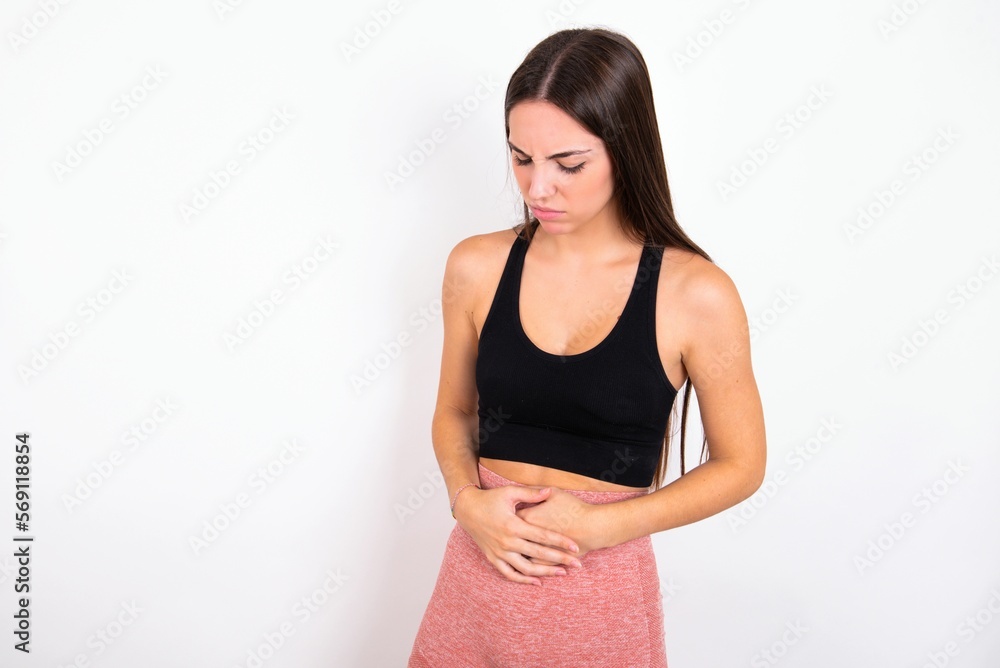 young woman wearing sportswear over white studio background suffering from strong stomachache. young woman wearing sportswear over white studio background suffering from strong stomachache.