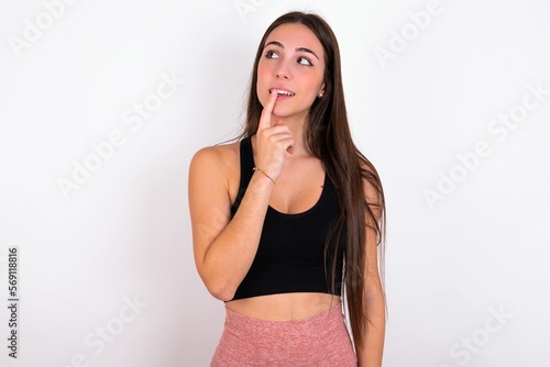 young woman wearing sportswear over white studio background with thoughtful expression, looks to the camera, keeps hand near face, bitting a finger thinks about something pleasant.
