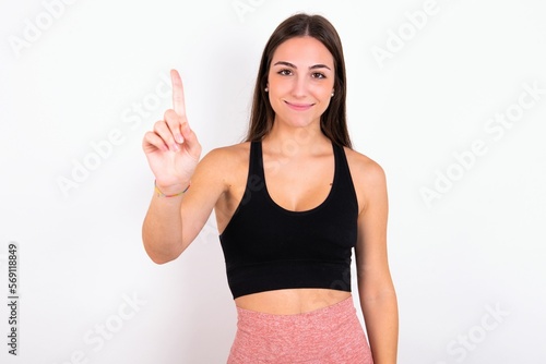 young woman wearing sportswear over white studio background showing and pointing up with fingers number one while smiling confident and happy. © Jihan