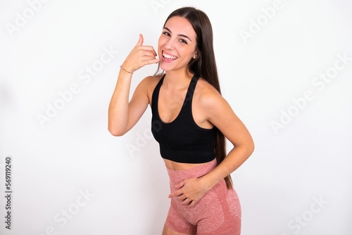 young woman wearing sportswear over white studio background imitates telephone conversation, makes phone call gesture with hands, has confident expression. Call me!