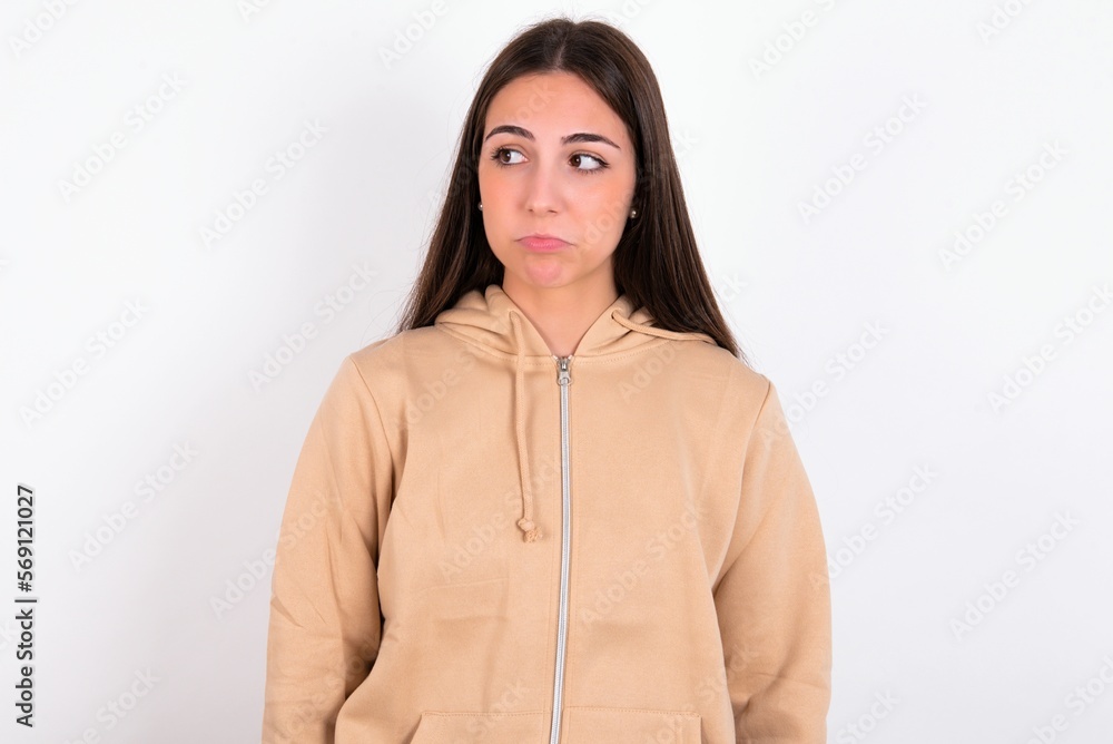Dissatisfied young woman wearing beige sweater over white studio background purses lips and has unhappy expression looks away stands offended. Depressed frustrated model. Dissatisfied young woman wearing beige sweater over white studio background purses lips and has unhappy expression looks away stands offended. Depressed frustrated model.