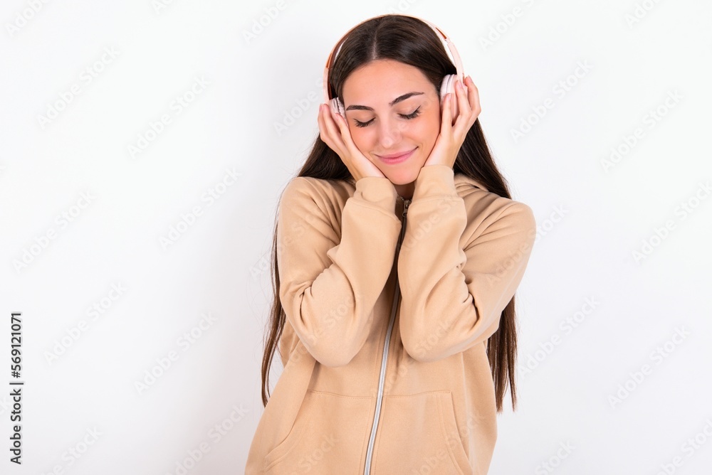 Pleased young woman wearing beige sweater over white studio background enjoys listening pleasant melody keeps hands on stereo headphones closes eyes. Spending free time with music Pleased young woman wearing beige sweater over white studio background enjoys listening pleasant melody keeps hands on stereo headphones closes eyes. Spending free time with music