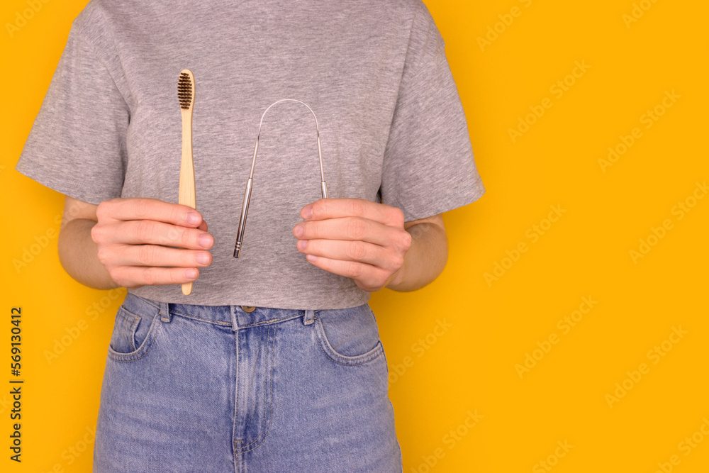 Female hands hold wooden toothbrush and metallic tongue scraper. Zero waste hygiene concept with copy space in front of yellow background. - obrazy, fototapety, plakaty 