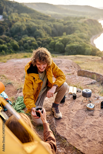 man giving cup of tea to female sitting in tent, at sunny ecening in autumn season. curly caucasian guy in yellow coat spend time with girlfriend in nature, mountains and river in the background photo