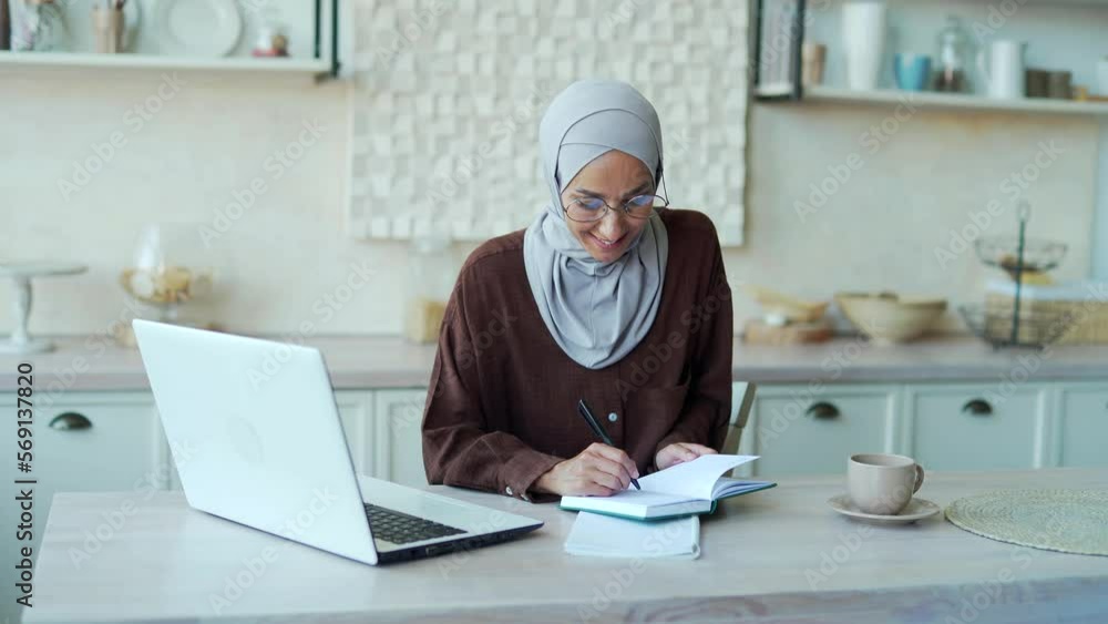 Smiling young Muslim woman in hijab student with glasses having remote lesson listening teacher lecturer making notes and looking at the laptop screen in room indoor Distance online education concept Smiling young Muslim woman in hijab student with glasses having remote lesson listening teacher lecturer making notes and looking at the laptop screen in room indoor Distance online education concept