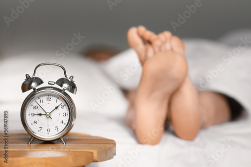 Feet of caucasian millennial lady lies on bed under white blanket, sleep at morning, focus on alarm clock on table