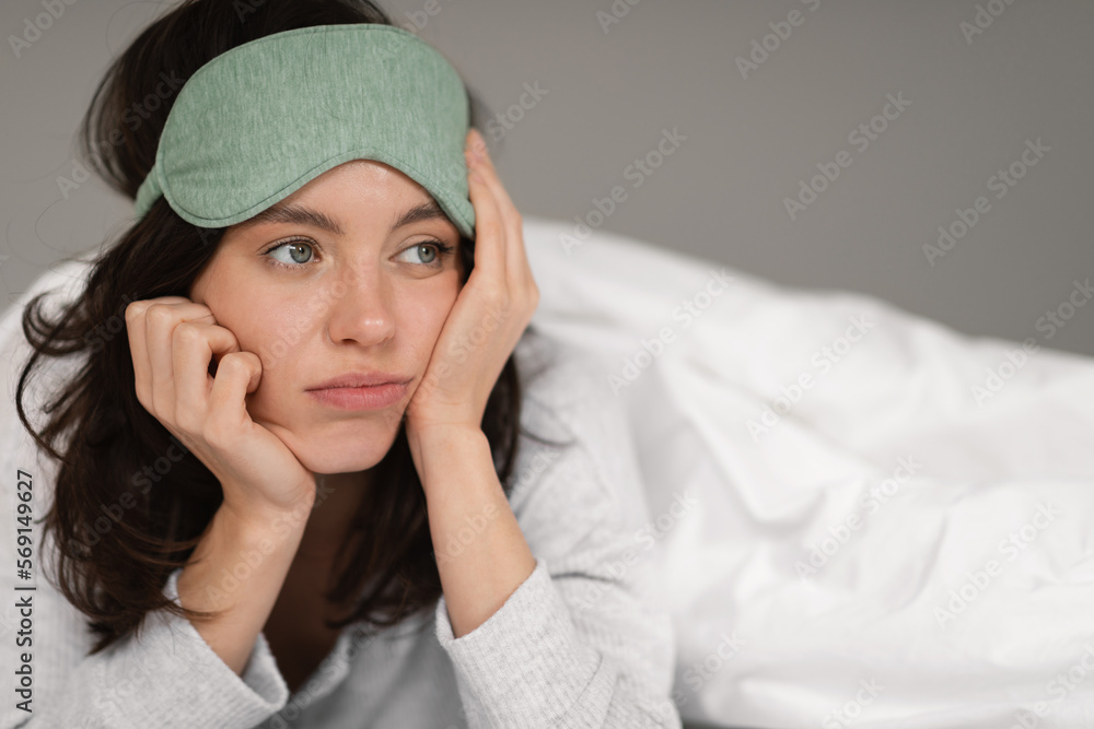 Unhappy tired caucasian millennial lady in sleep mask lies on bed under white blanket, looks at empty space Unhappy tired caucasian millennial lady in sleep mask lies on bed under white blanket, looks at empty space