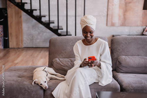 Smiley African businesswoman in white dress and turban sitting on cozy couch with white dog talking by phone. Purposeful African American woman makes call from hotel room. Owner and pet at home.