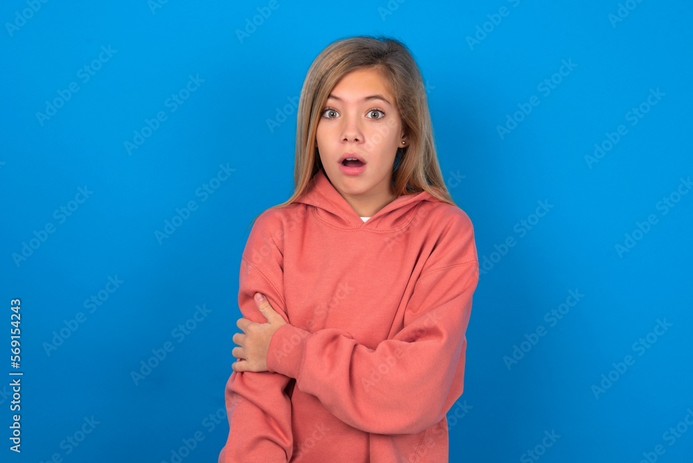 Fototapeta premium Shocked embarrassed caucasian teenager girl wearing pink sweater over blue background keeps mouth widely opened. Hears unbelievable novelty stares in stupor