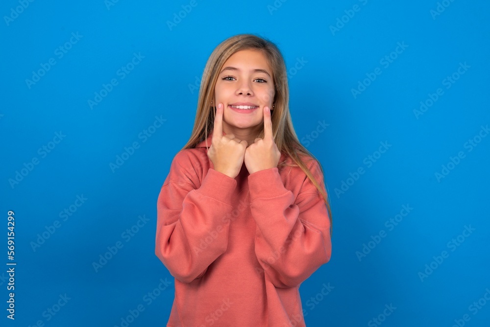 Happy caucasian teenager girl wearing pink sweater over blue background with toothy smile, keeps index fingers near mouth, fingers pointing and forcing cheerful smile Happy caucasian teenager girl wearing pink sweater over blue background with toothy smile, keeps index fingers near mouth, fingers pointing and forcing cheerful smile