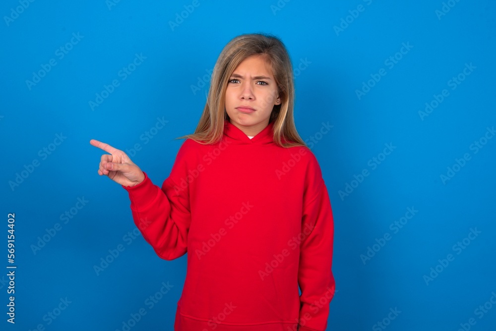 Fototapeta premium Serious caucasian teenager girl wearing red sweater over blue background smirks face points away on copy space shows something unpleasant. Look at this advertisement. Big price concept