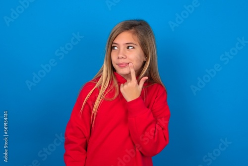 Lovely dreamy caucasian teenager girl wearing red sweater over blue background keeps finger near lips looks aside copy space.