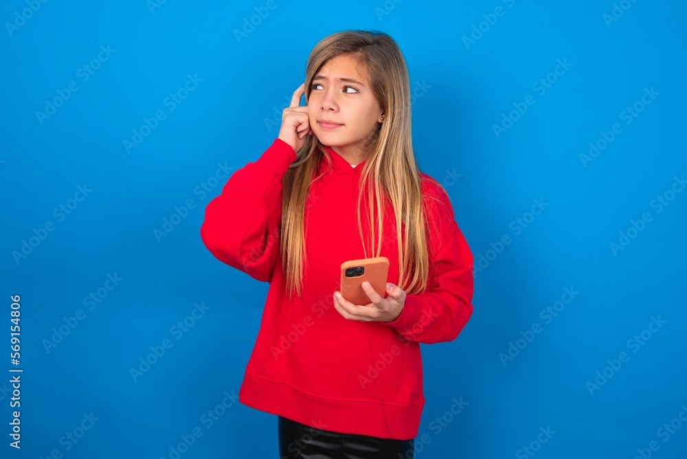 Pensive caucasian teenager girl wearing red sweater over blue background keeps index finger on temple, holds modern smartphone and concentrated aside Pensive caucasian teenager girl wearing red sweater over blue background keeps index finger on temple, holds modern smartphone and concentrated aside