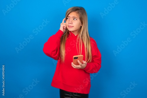 Pensive caucasian teenager girl wearing red sweater over blue background keeps index finger on temple, holds modern smartphone and concentrated aside © Roquillo