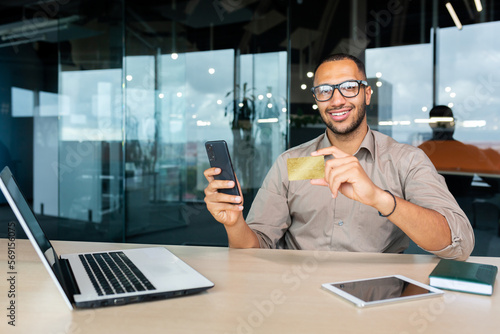 Portrait of successful African American businessman inside office, man smiling and looking at camera, worker holding phone and bank credit card working at work using laptop.