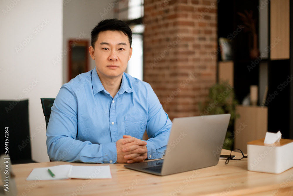 Serious middle aged asian male entrepreneur working on laptop and looking at camera, sitting at workplace at home office Serious middle aged asian male entrepreneur working on laptop and looking at camera, sitting at workplace at home office