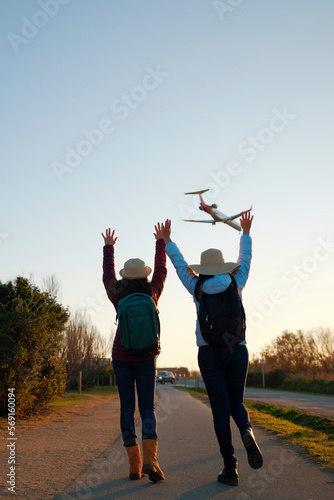 Back view of two female travelers with backpacks raising their arms and watching the planes go by.