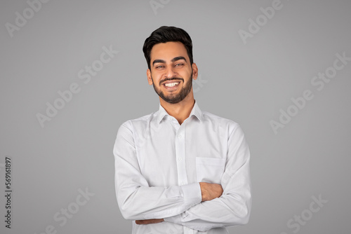 Smiling Handsome Young Arab Businessman With Folded Arms Standing Over Grey Background © Prostock-studio