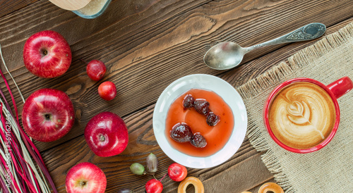cup of cappuccino coffee, ripe apples, actinidia berries, bagels, strawberry jam in white saucer, spoon, red napkin at white polka dots on wooden table with decorative colorful  dry straw. top view