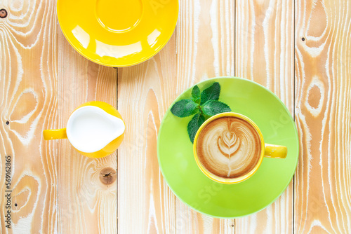 cup of cappuccino coffee on  light green plate and yellow milk jug .on wooden table, top view