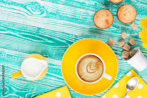 cup of cappuccino coffee on yellow plate and yellow milk jug cane sugar,  cakes, dandelions, teaspoon on turquoise colored wooden table with yellow napkin at polka dots top view