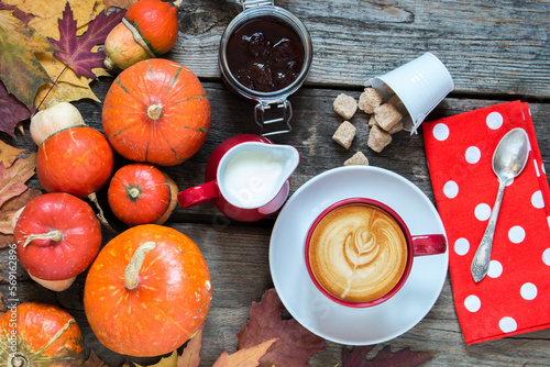 cup of cappuccino coffee milk jug, strawberry jam, red napkin at polka dots on wooden table decorated by falling leaves and pumpkins, top view