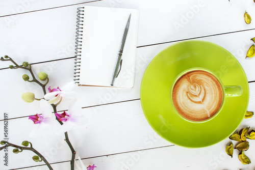 cappuccino coffee in green cup,  orchid flower, notebook and silver pen,  green dry flower decor scattered on white painted wooden table, top view