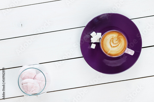 cup of cappuccino coffee on violet plate with sugar,  marshmallows in a glass vase on white colored wooden table , top view