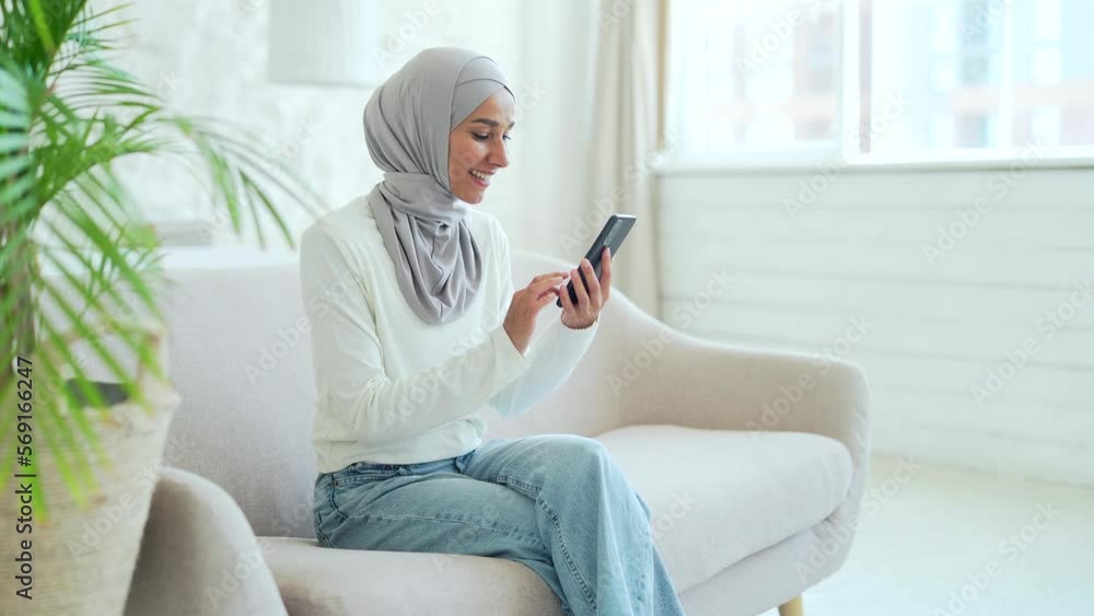 Smiling Muslim woman in hijab hold smartphone scrolling and watching social media in living room at home Young female browsing products in online internet store using phone texting chatting indoors Smiling Muslim woman in hijab hold smartphone scrolling and watching social media in living room at home Young female browsing products in online internet store using phone texting chatting indoors