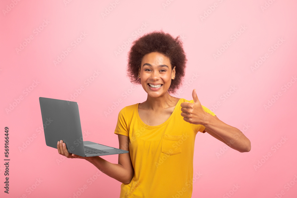 Glad young african american curly female in yellow t-shirt with laptop show thumb up isolated on pink background Glad young african american curly female in yellow t-shirt with laptop show thumb up isolated on pink background