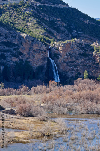 Beautiful landscape of vertical domeño waterfall flowing over the mountain rock with dry bushes and turia river in the foreground, near domeño old town in Valencia, Spain photo