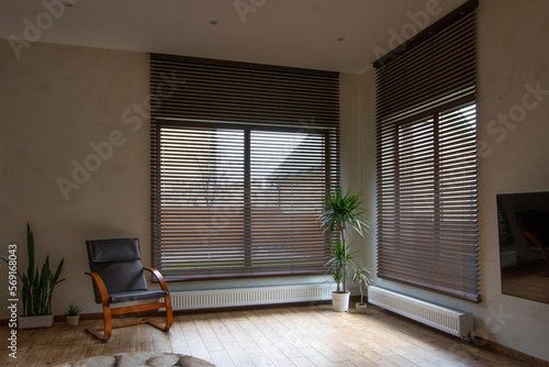 Wooden blinds on large windows in the interior. Living room with armchair and houseplants near windows with wood blinds. Motorized jalousie in the smart house. photo