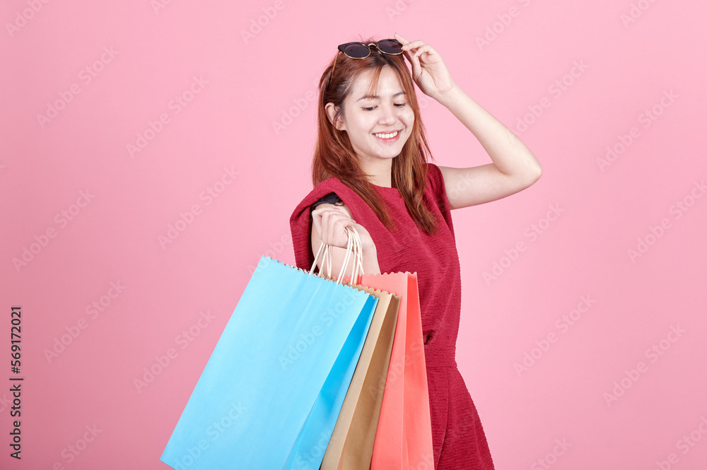 Portrait of an excited asian girl wearing red dress and sunglasses holding shopping bags isolated over pink background. Asia woman carrying colorful bags shopping.