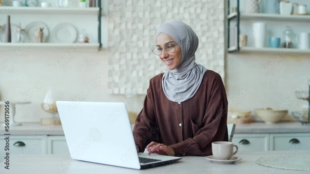Smiling Muslim woman in hijab enjoying cup of coffee or tea and continue her work typing on laptop in light room Attractive female doing job browsing web at kitchen Online studying or work from home Smiling Muslim woman in hijab enjoying cup of coffee or tea and continue her work typing on laptop in light room Attractive female doing job browsing web at kitchen Online studying or work from home