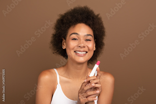 Cheerful young african american curly female with perfect skin in white top uses moisturizing lipstick for lips