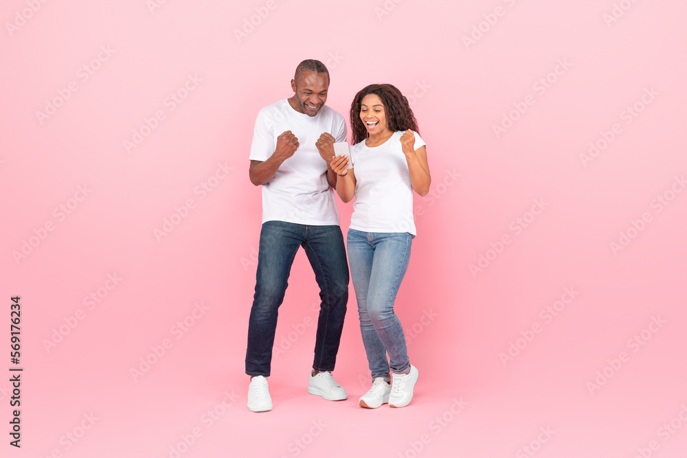 Cheerful black couple gambling together online using smartphone, rasising hands up and smiling, pink studio background Cheerful black couple gambling together online using smartphone, rasising hands up and smiling, pink studio background