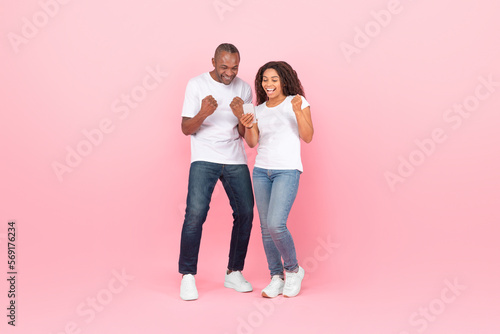 Cheerful black couple gambling together online using smartphone, rasising hands up and smiling, pink studio background